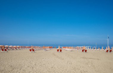 Açık mavi gökyüzü olan güneşli bir günde turuncu ve beyaz şemsiyeli kırmızı plaj sandalyelerinin manzarası..