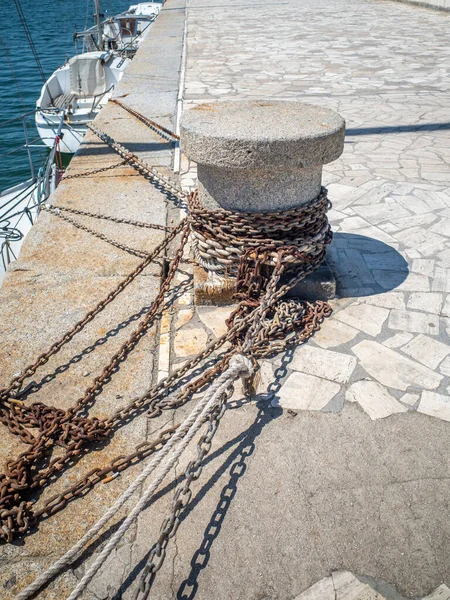 A concrete capstan on a stone quayside with chains and ropes attached on a sunny day.