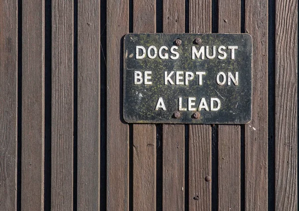 木墙上的黑白相间的金属标志上写着狗必须保持领先 — 图库照片