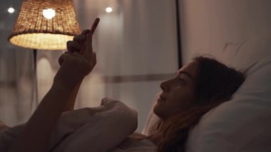 Genç bir kadın gece yatakta uzanıyor ve akıllı telefonunda geziniyor.