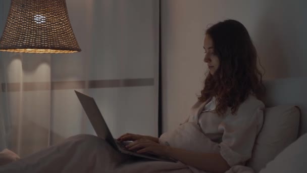 Mujer joven concentrada que trabaja por la noche en su dormitorio usando computadora portátil y escribiendo mensaje — Vídeos de Stock