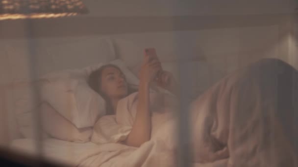 Gluurt als een meisje ligt in bed en leest uit de telefoon — Stockvideo