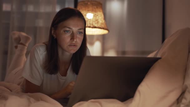 Mujer joven concentrada que trabaja por la noche en su dormitorio usando computadora portátil y escribiendo mensaje — Vídeos de Stock