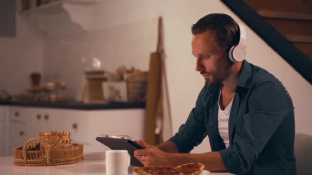 Ο άνθρωπος που εργάζεται τη νύχτα στην κουζίνα του χρησιμοποιώντας tablet και να ακούσετε μουσική με ακουστικά — Αρχείο Βίντεο
