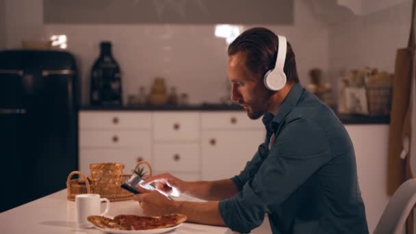 Homem trabalhando à noite em sua cozinha usando tablet e ouvir música com fones de ouvido — Vídeo de Stock