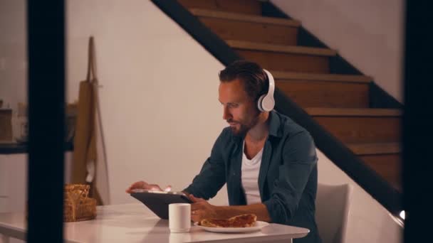Muž pracující v noci ve své kuchyni pomocí tabletu a poslouchat hudbu se sluchátky — Stock video