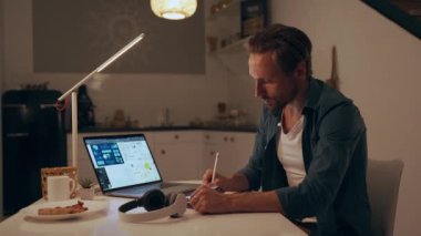 Geceleri mutfakta tablet kullanan ve kulaklıkla müzik dinleyen bir adam.