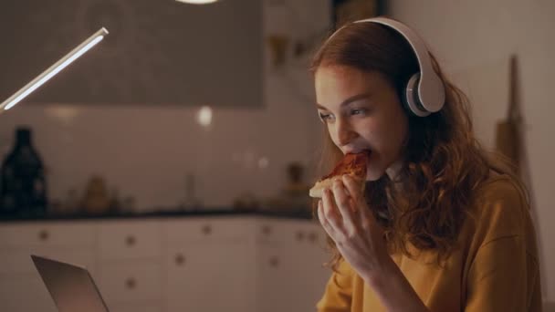 女性は夜仕事をしたり、ピザを食べたり、ノートパソコンに入力したり、音楽を聴いたりします。 — ストック動画