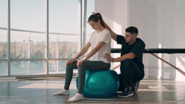 Mujer joven con su fisioterapeuta durante el entrenamiento de rehabilitación en la pelota de gimnasia en la clínica — Vídeos de Stock