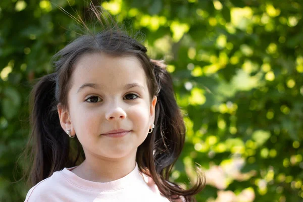 Atkuyruklu neşeli küçük kız, gülümsüyor. Önüne, yeşil yapraklı bir arka plana bakıyor.