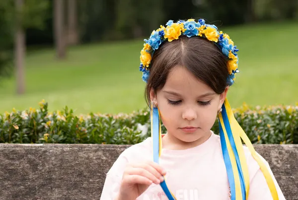 头上戴着乌克兰民间装饰品的可怜小女孩 小孩子坐在户外的长椅上 触摸花环缎带 — 图库照片