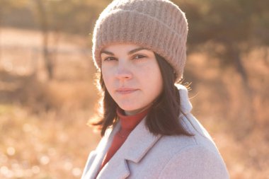 Örgü şapkalı ciddi bir kız, sonbahar soluk bir gölet manzarasının zeminine karşı bir fonda ve bir paltoda duruyor. Model doğrudan kameraya bakar.