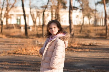 Kabarık kapüşonlu ceketli yalnız üzgün bir kız dönüp kameraya bakıyor. Soğuk bir sonbahar sokağında, sıcak elbiseli bir çocuk.