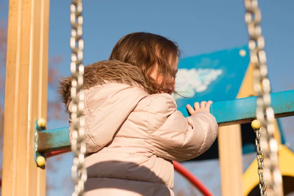 Küçük bir çocuk, uzun kızıl saçlı, arka planda mavi bir gökyüzü, oyun alanında sıcak giysiler içinde.