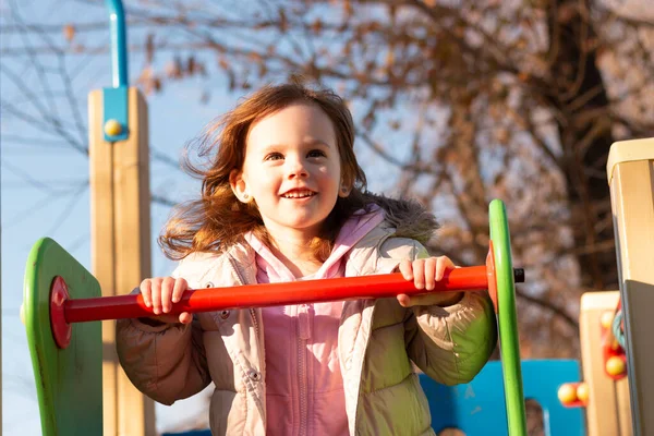 Küçük bir çocuk, uzun kızıl saçlı, arka planda mavi bir gökyüzü, oyun alanında sıcak giysiler içinde.