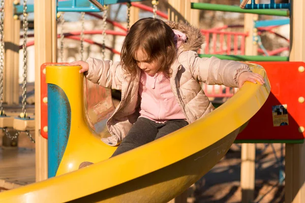 Küçük bir çocuk, uzun kızıl saçlı, arka planda mavi bir gökyüzü, oyun alanında sıcak giysiler içinde.