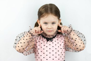 Beyaz arka planda benekli iki at kuyruklu gülümseyen küçük kız parmaklarıyla kulaklarını kapatıyor. Stüdyo fotoğrafı