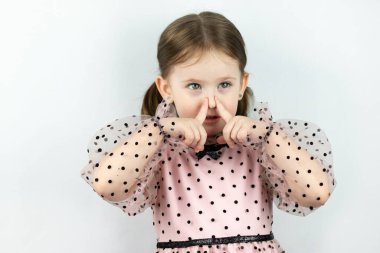 Beyaz arka planda benekli iki at kuyruklu gülümseyen küçük kız parmaklarıyla burnunu kapatıyor. Stüdyo fotoğrafı
