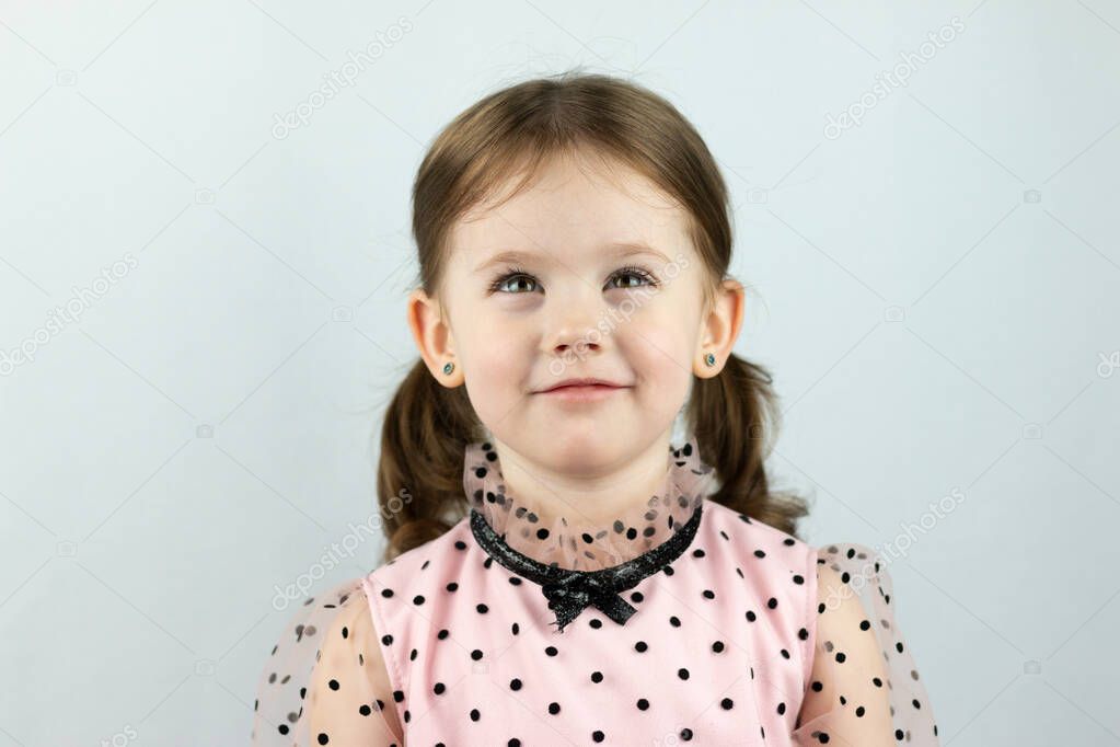 Smiling little girl with two ponytails in a dress with polka dots on a white background dreamily looks up. Studio photo