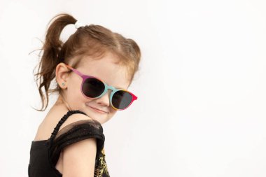 A little girl with two ponytails and a black and gold dress looks playfully at the camera. Studio photo on a white background of a child in sunglasses