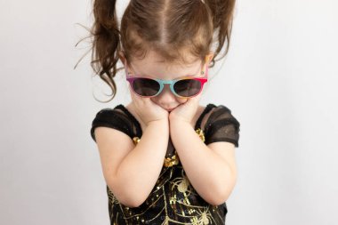A little sad girl in sunglasses with two ponytails and a black and gold dress holds her head down with her hands. Studio photo of a child on a white background