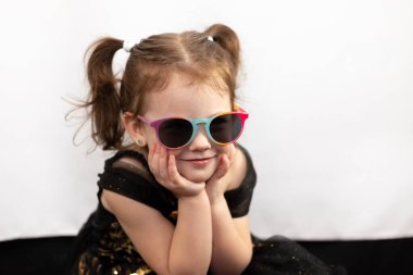 Beyaz arka planda şirin bir bebeğin stüdyo fotoğrafı. İki at kuyruklu, güneş gözlüklü, siyah ve altın rengi elbiseli, neşeli bir kız başını avuçlarına koydu.