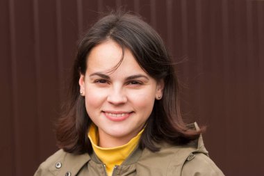 Gayri resmi ceketli, siyah saçlı, kahverengi arka planda duran güzel, sevimli bir kız.