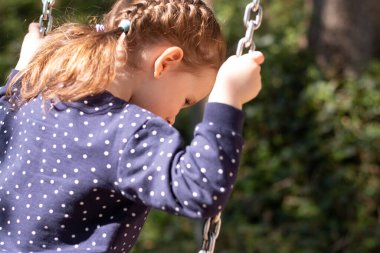 Küçük gülümseyen kız, arka planda salıncakta sallanırken yere bakar.