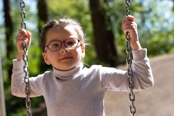 Piccola Ragazza Carina Occhiali Sorridente Cavalca Altalena Parco Giochi Nel Foto Stock