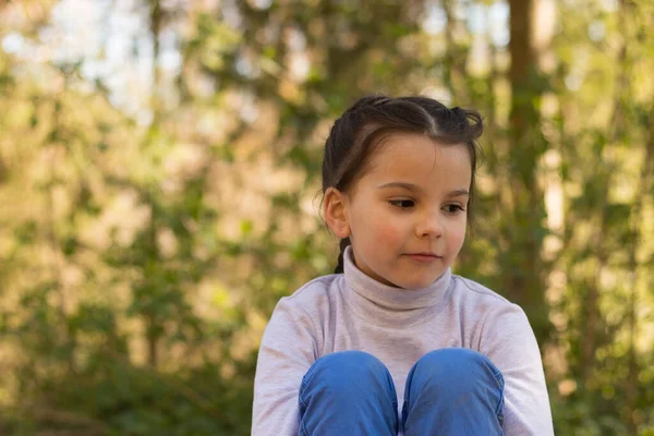 Balıkçı yaka kazak ve kot pantolon giymiş, doğal bir geçmişi olan, gülümseyen küçük bir kız öğrenci yere oturmuş bacaklarına sarılıyor ve gözlerini kaçırıyor.