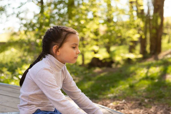 Balıkçı yaka kazak ve kot pantolon giymiş, doğal bir geçmişi olan, gülümseyen küçük bir kız öğrenci yere oturmuş bacaklarına sarılıyor ve gözlerini kaçırıyor.