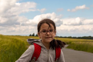 Gözlüklü küçük bir kız gülümsüyor kırsal bir manzaranın arka planında duruyor. Okul kızı tarlanın ortasında yolda.