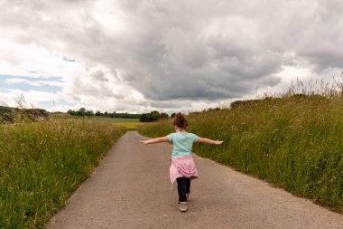 Küçük bir çocuk yol boyunca bir tarlanın ortasında kollarını açmış koşuyor.