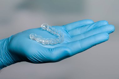 Diş hekimi mavi eldivenlerle dişçi odasının önünde şeffaf, çıkarılabilir bir sıralayıcı sunuyor.
