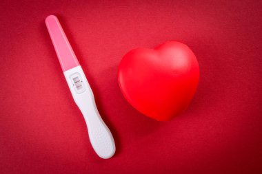 Kırmızı arka planda pozitif hamilelik testi ve plastik kalp. Yeni yaşam konsepti