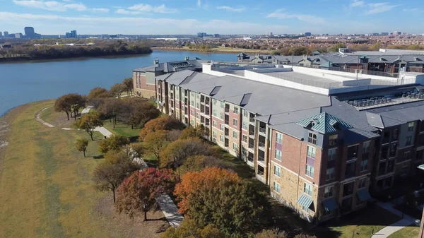 Aerial view riverside apartment complex wit downtown Las Colinas, Irving, Texas in background. Multistory rental complex with concrete pathway sidewalk, colorful fall foliage, sunny blue sky