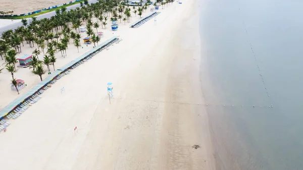 Long white sandy shoreline with row of folding chairs, palm trees and tourist activity at Tuan Chau, Ha Long Bay, Quang Ninh. Top view tropical beach travel and recreational destination in Vietnam