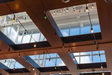Lookup view of colorful light bulb string on ceiling near glass roof and wooden exposure beam structure of terminal airport bar in Houston, Texas, USA. Modern design interior, furniture background