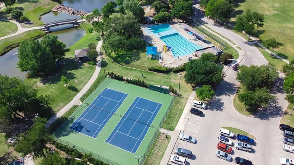 Aerial view large tennis court and swimming pool at community recreational center with trails and pond near Dallas, Texas, USA. Green park surrounded by mature trees, busy parking lots and outdoors