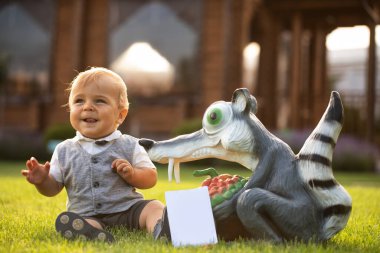 A cheerful baby in the backyard is sitting on a bright juicy green lawn and playing with garden toys. Selective focus.
