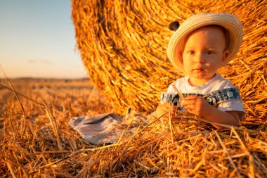 A small boy in a hat plays with straw near a large round straw bale. Sunset. Active summer vacation in the countryside.