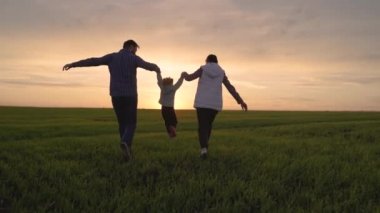 Baba kızı anne, memeler, mutlu bir çocuk ebeveynlerinin elini tutuyor, bir çocuk yeşil çimlerin üzerinde zıplıyor. Aile baharda gün batımında parkta yürüyüş, sağlıklı bir çocukluk. Aile hafta sonu sahada