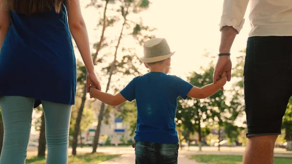 Vacaciones familiares, fin de semana. Padres felices y niños caminando en el parque. Feliz familia camina con un niño en la calle tranquila en verano al atardecer. Papá, hijo, mamá y tomar las manos en la acera en la ciudad en el sol. — Foto de Stock