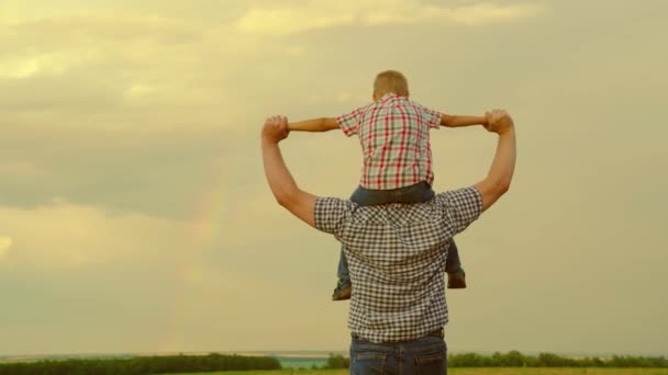 Pai e criança brincam, fantasiam. Família feliz descansando no parque na frente do arco-íris no céu. Criança feliz, filho senta-se no pescoço de seus pais, joga piloto, voa como super-herói, sonha em voar com seu pai. — Vídeo de Stock