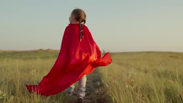 Šťastná superhrdinka běhající trávou na poli v červeném plášti, plášť třepetající se ve větru. Dítě sní, že se stane superhrdinou. Děvče v červeném plášti snového výrazu. Dětské hry sny Dovolená — Stock video