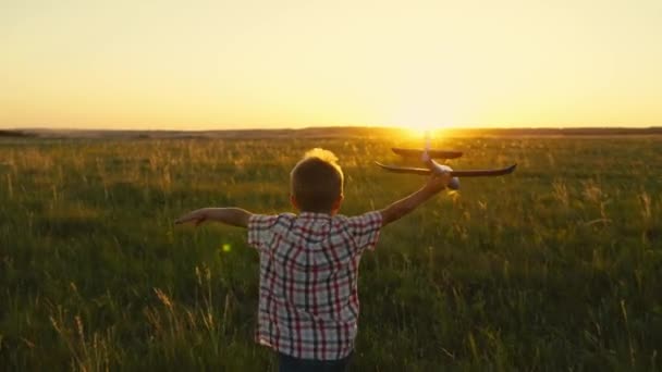 Kind jongen rent met speelgoed vliegtuig op het veld bij zonsondergang. Gelukkig kind loopt in het park, spelen met speelgoed vliegtuig in de zomer in de natuur. Gelukkige familie. Jongen droomt van vliegen. Zorgeloos buiten spelend kind — Stockvideo
