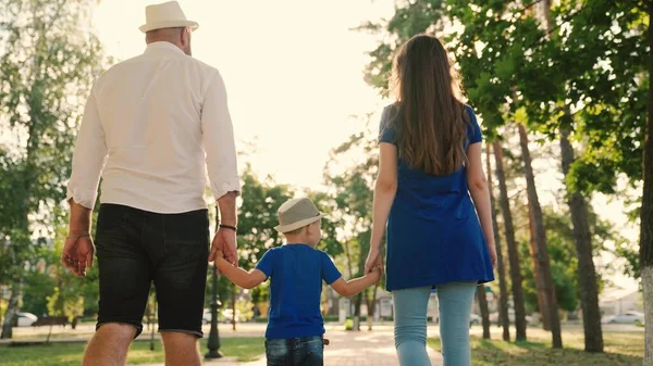 Feliz familia camina con un niño en la calle tranquila en verano al atardecer. Vacaciones familiares, fin de semana. Padres felices y niños caminando en el parque. Papá, hijo, mamá y tomar las manos en la acera en la ciudad en el sol. — Foto de Stock