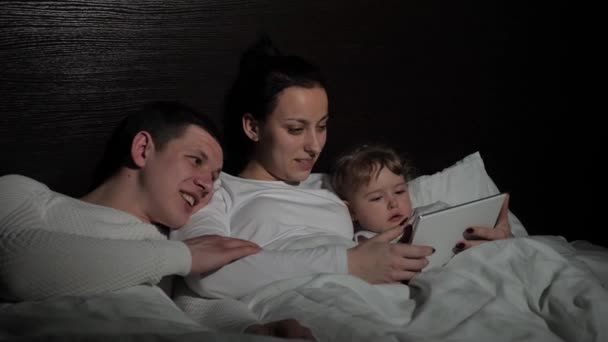 Lille barn, datter og forældre ser tegneserie online ved hjælp af digital tablet, før de går i seng på sengen. Lykkelig familie hviler sammen om aftenen derhjemme med tablet. Online familieuddannelse – Stock-video