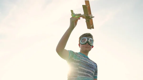 Criança despreocupada em óculos piloto brinca com avião de brinquedo ao ar livre. Menino feliz brincando com brinquedo avião de madeira na frente do céu. O miúdo tem um avião de brinquedo na mão. Família feliz jogando no parque — Fotografia de Stock