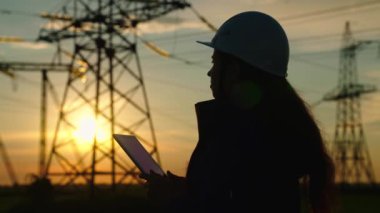 İnşaat mühendisi, kadın elektrik tedarikçisi açık havada çalışıyor. Çevre dostu elektrik. Modern teknolojiler. Güvenlik kaskındaki güç mühendisi güç hattını ve dijital tableti kontrol ediyor.